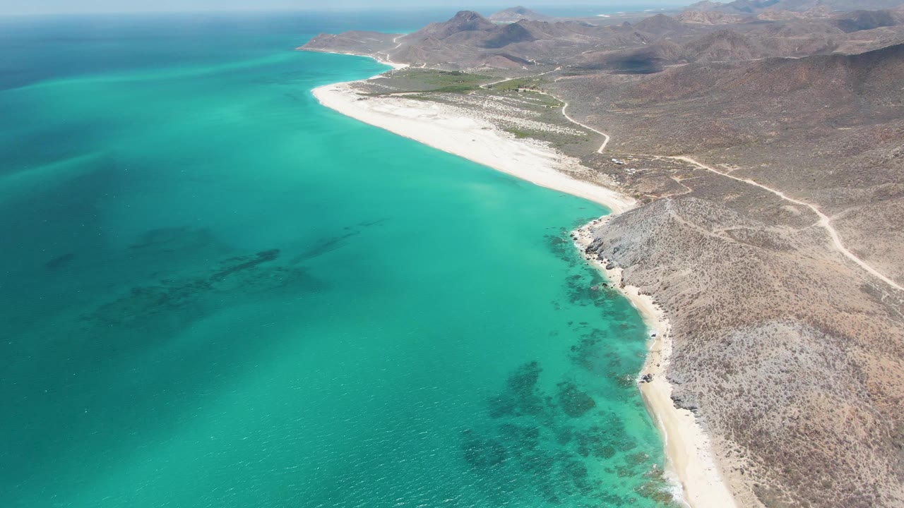 Aerial view at Barakas Beach with turquoise shallows filling the left side, a narrow white beach running through the center, rocky coastal ground at right, mountains beyond, and no visible people.