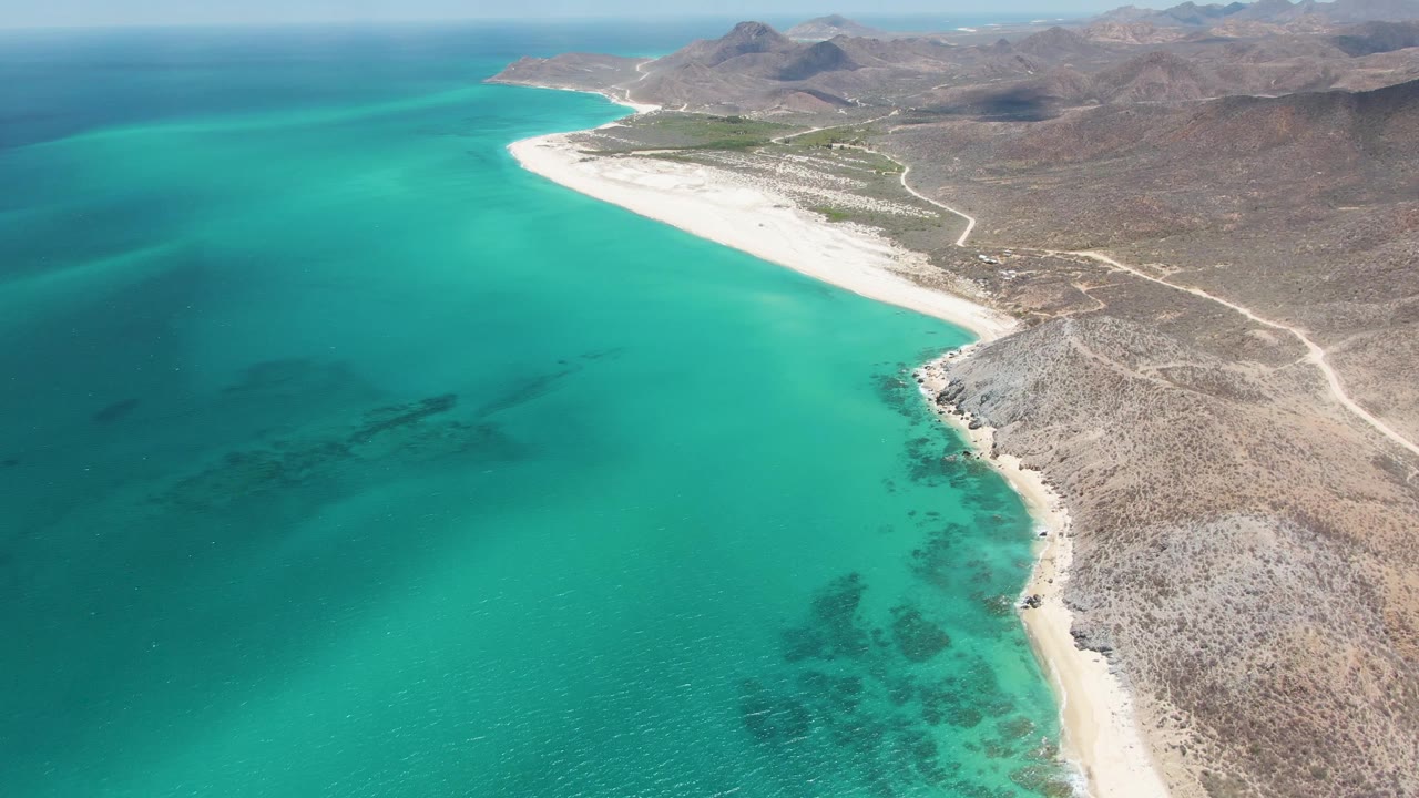 Mid-clip aerial frame over Barakas Beach with a broad turquoise bay on the left, a bright white sand strip along the coast, dry rocky land and pale tracks on the right, and no visible people.