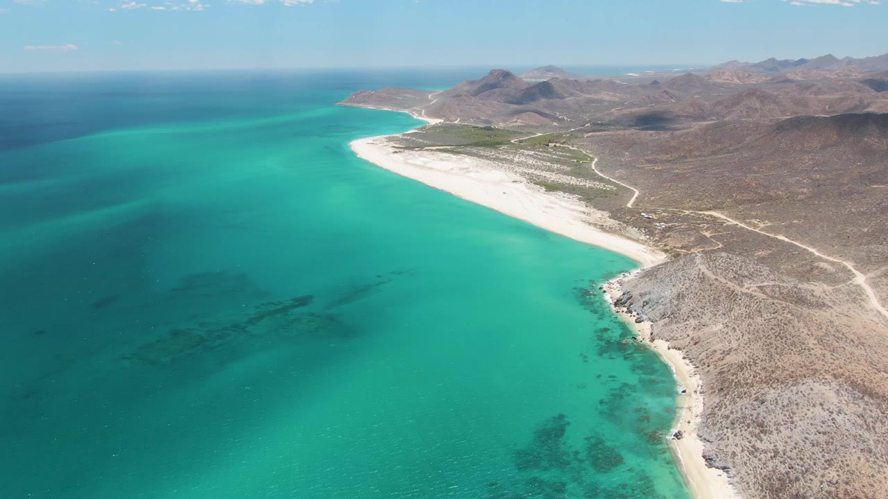 Aerial view over Barakas Beach in Baja California Sur, with turquoise water, a pale curving shoreline, rocky desert land, distant mountains, and no visible people.