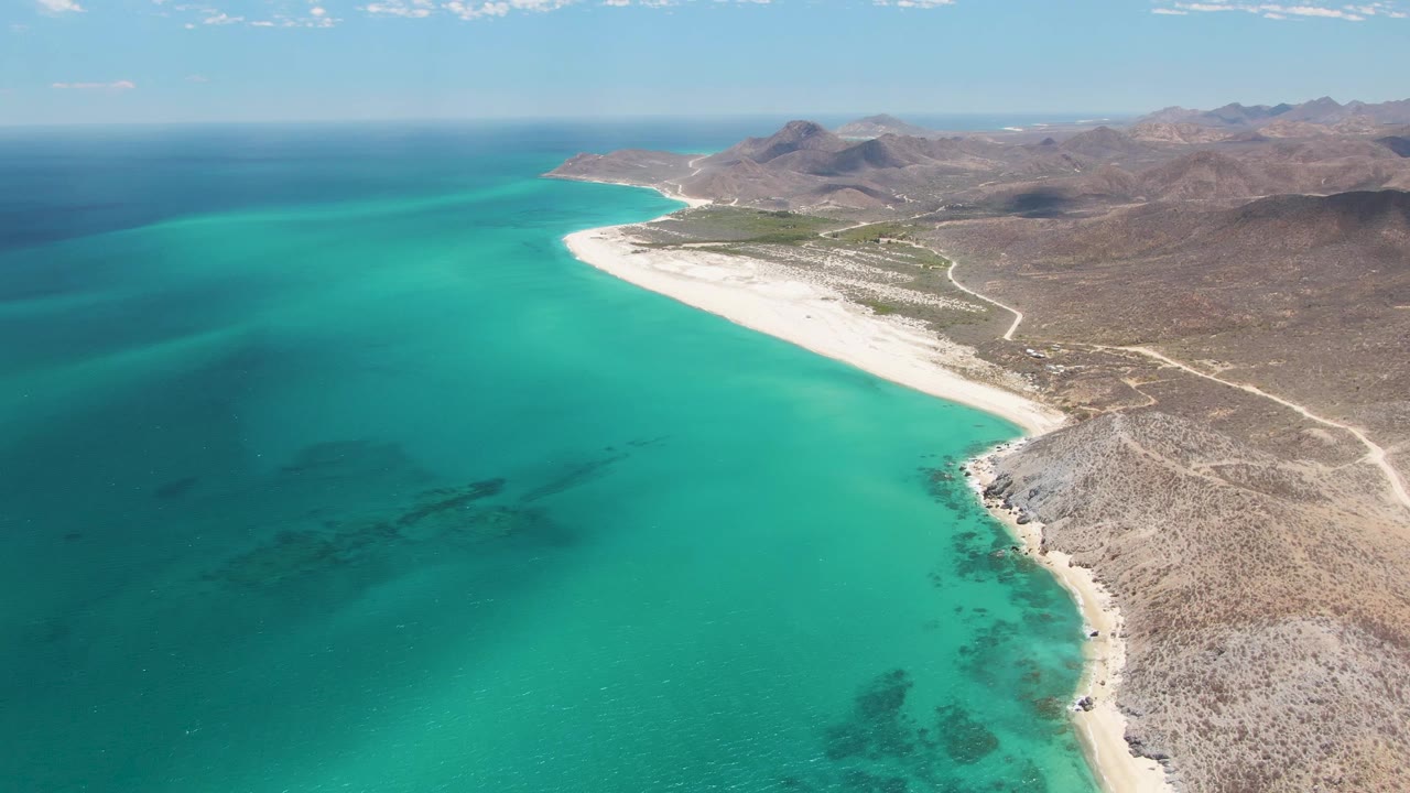 Aerial frame of Barakas Beach showing the blue-green water spread across the left side, the white beach curving along the coast, dry Baja California Sur terrain, and no visible people.