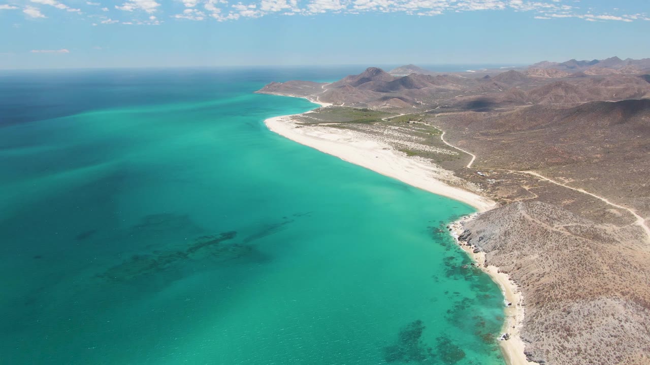 Wide drone-like view of Barakas Beach with more open turquoise water in the foreground, the beach running diagonally toward the mountains, and no visible people on the sand or land.