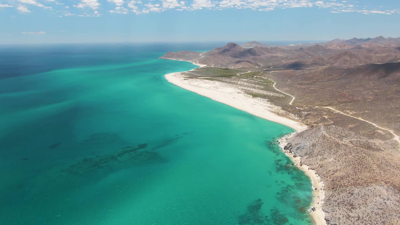 Aerial view of Barakas Beach at mid-clip, with teal water filling the left foreground, a pale beach arc through the center, dry rocky land to the right, mountains on the horizon, and no visible people.