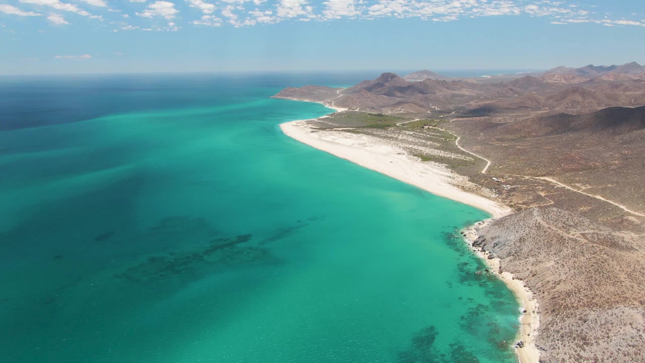 Second aerial moment at Barakas Beach with blue-green water spread across the foreground, white sand tracing the coast, brown hills inland, and no visible people.