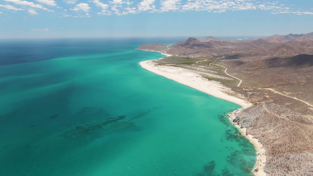 Mid-clip aerial frame at Barakas Beach with calm turquoise water filling the lower left, the white beach running diagonally, rocky land to the right, and no visible people.