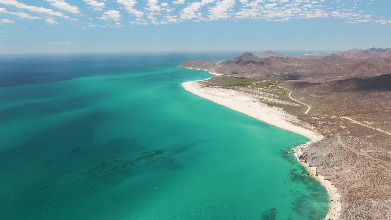 Wide aerial frame at Barakas Beach showing a broad turquoise bay, a pale sandy shoreline, dry rocky land, mountain ridges in the background, scattered clouds, and no visible people.