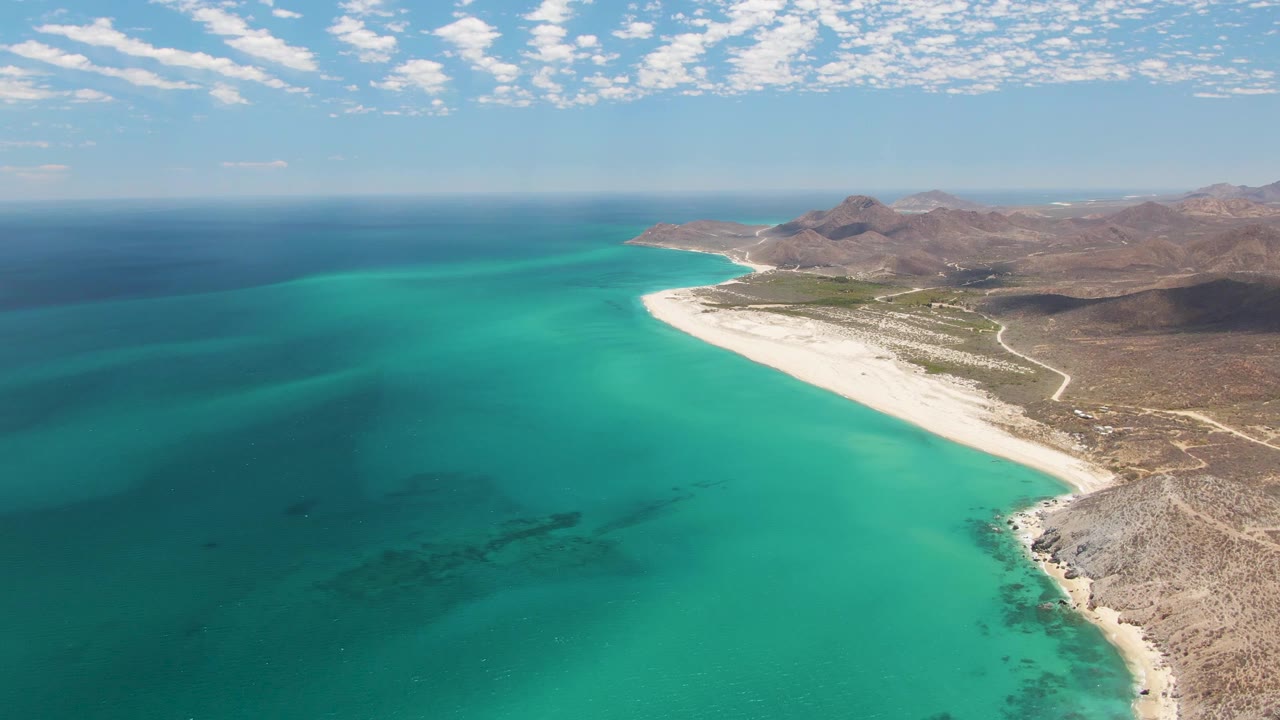 Aerial view of Barakas Beach in Baja California Sur with turquoise water on the left, a white sand shoreline curving toward rocky hills, and no visible people.