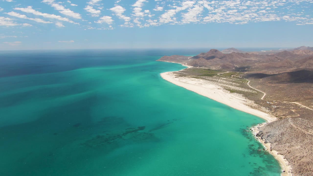 Aerial frame over Barakas Beach showing darker offshore water bands, bright shallow turquoise water, a pale sand curve, inland hills, and no visible people.