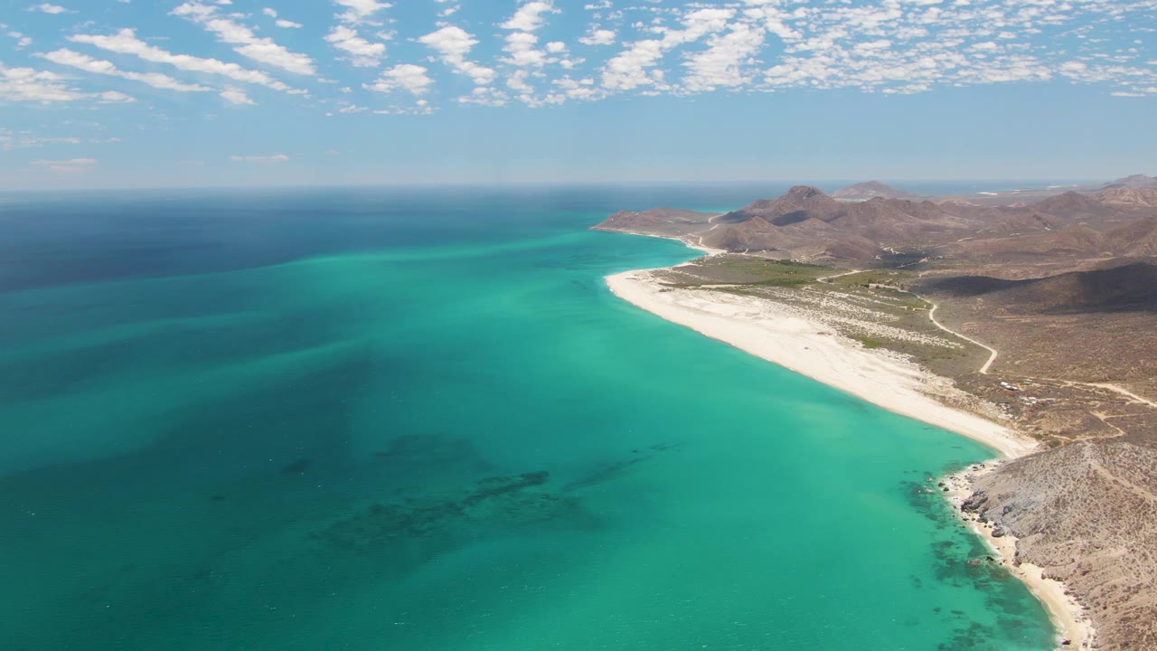 Aerial view of Barakas Beach with the shoreline running diagonally through the frame, turquoise ocean filling the left side, rugged brown hills to the right, and no visible people.