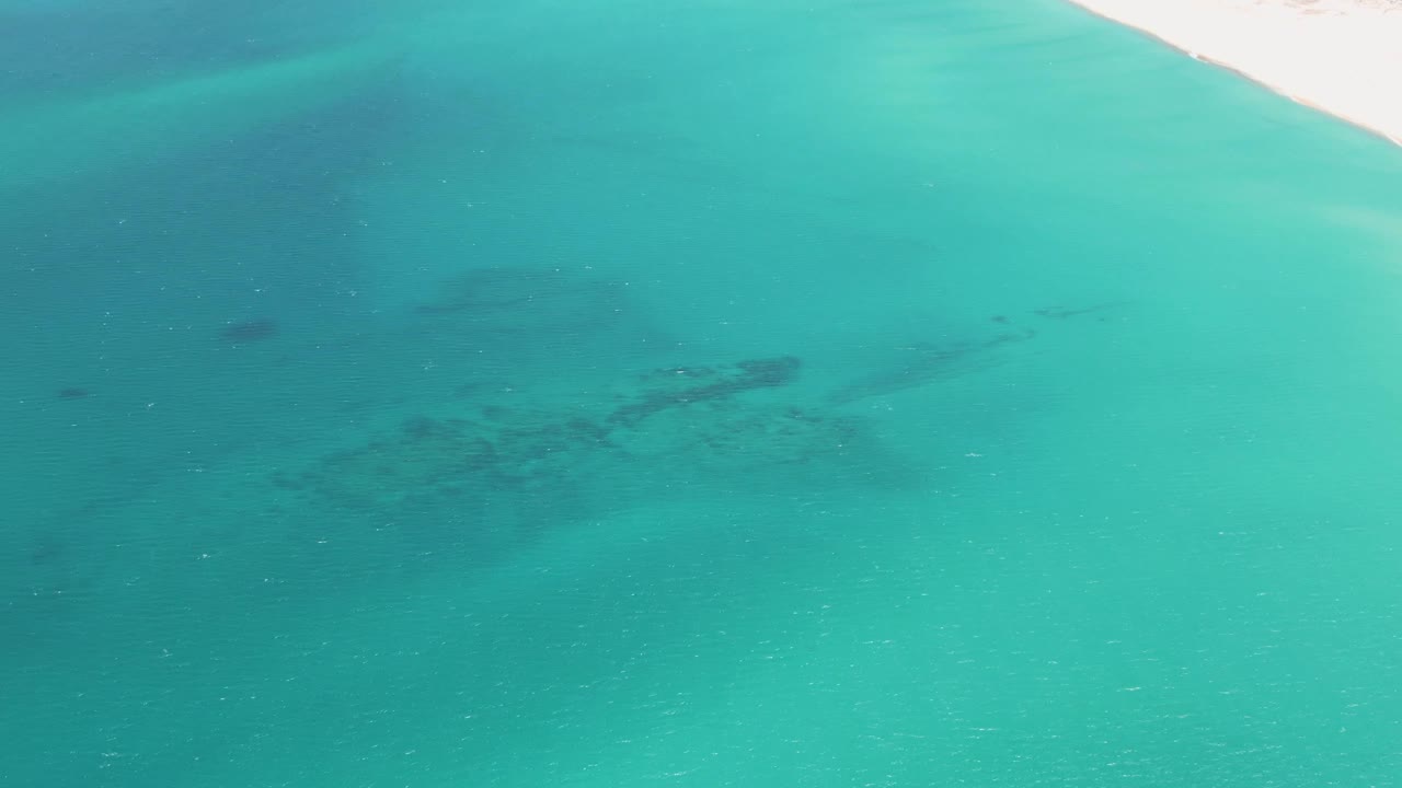 Aerial view over turquoise water at Barakas Beach, Baja California Sur, with a narrow strip of pale sand at the top edge and no visible people.