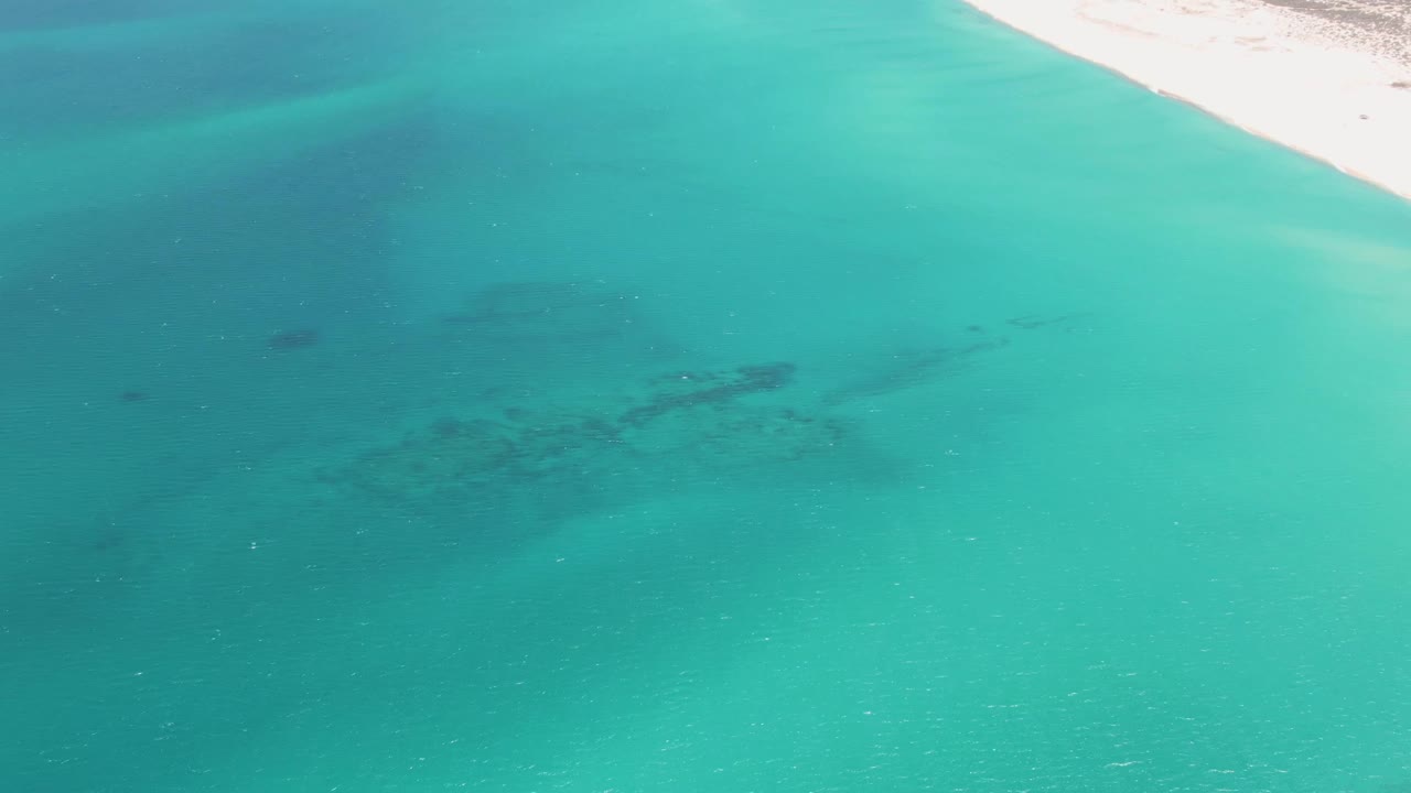 Aerial frame at Barakas Beach showing clear turquoise shallows, darker underwater patches, and a pale sandy beach entering from the upper right with no visible people.