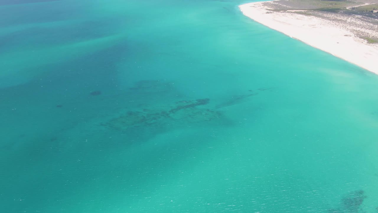 Aerial view of Barakas Beach with turquoise water across the frame, a pale shoreline at upper right, dry land beyond it, and no visible people.