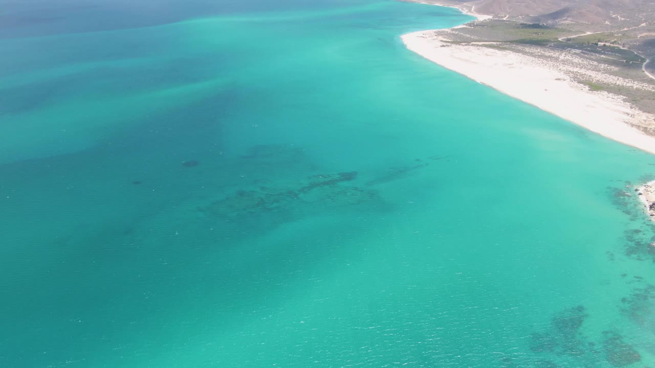 Wide aerial frame at Barakas Beach showing turquoise water in the foreground, a long pale beach, arid hills, a shoreline road, and no visible people.