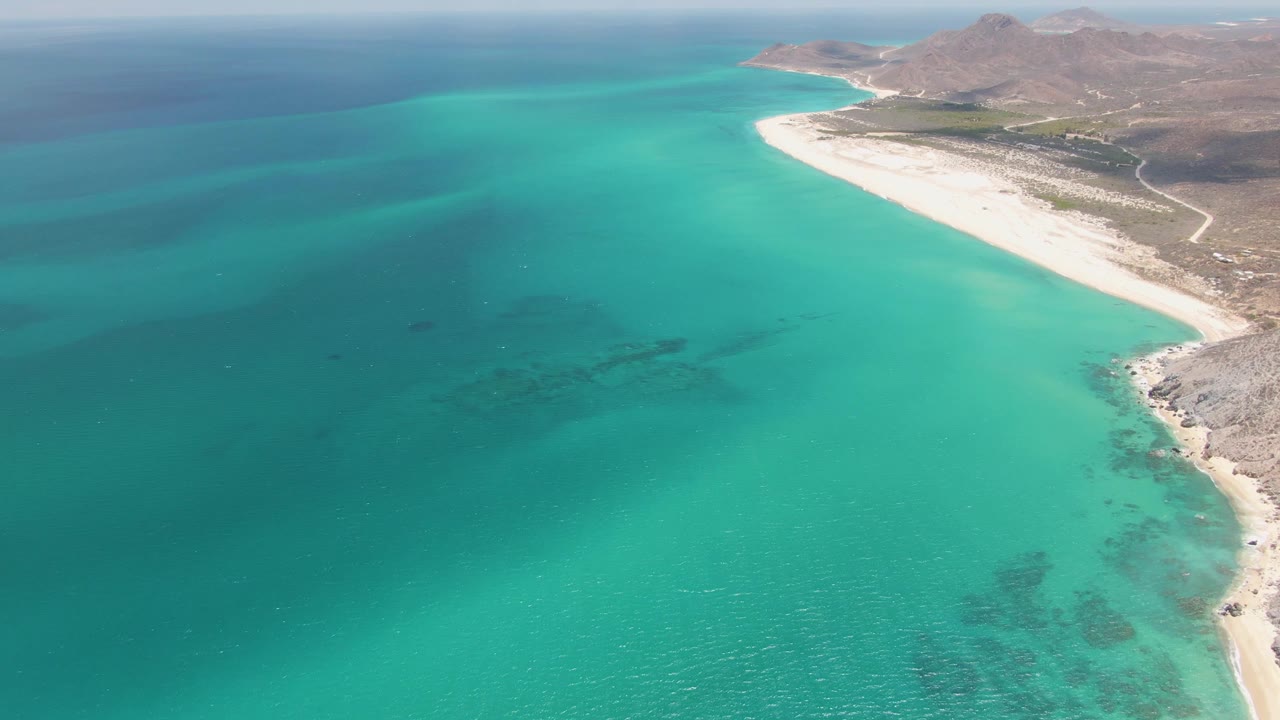 Opening aerial frame over Barakas Beach, Baja California Sur, showing turquoise coastal water, pale sand, rocky hills, a shoreline road, and no visible people.