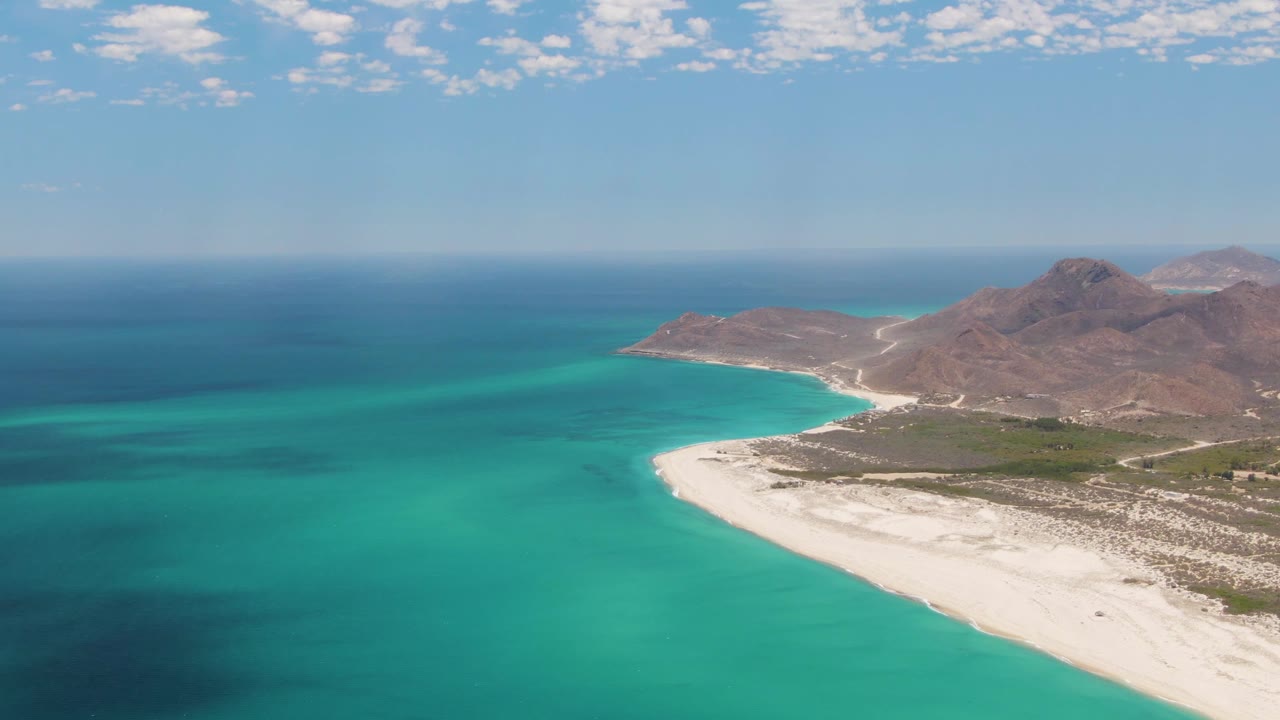 Aerial view of turquoise ocean beside a white sand beach and rocky mountains.