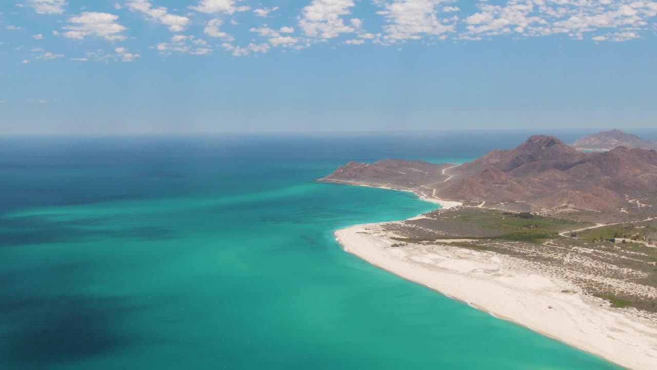 Aerial frame at Barakas Beach showing more open turquoise water on the left, the white beach sweeping through the right side, dry brown mountains behind it, and no visible people.