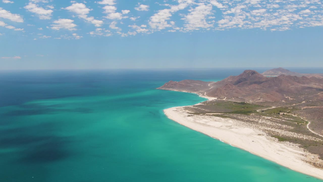 Mid-clip aerial view at Barakas Beach, Baja California Sur, with turquoise sea filling the lower frame, white sand and scrubland along the right, rocky hills behind, and no visible people.