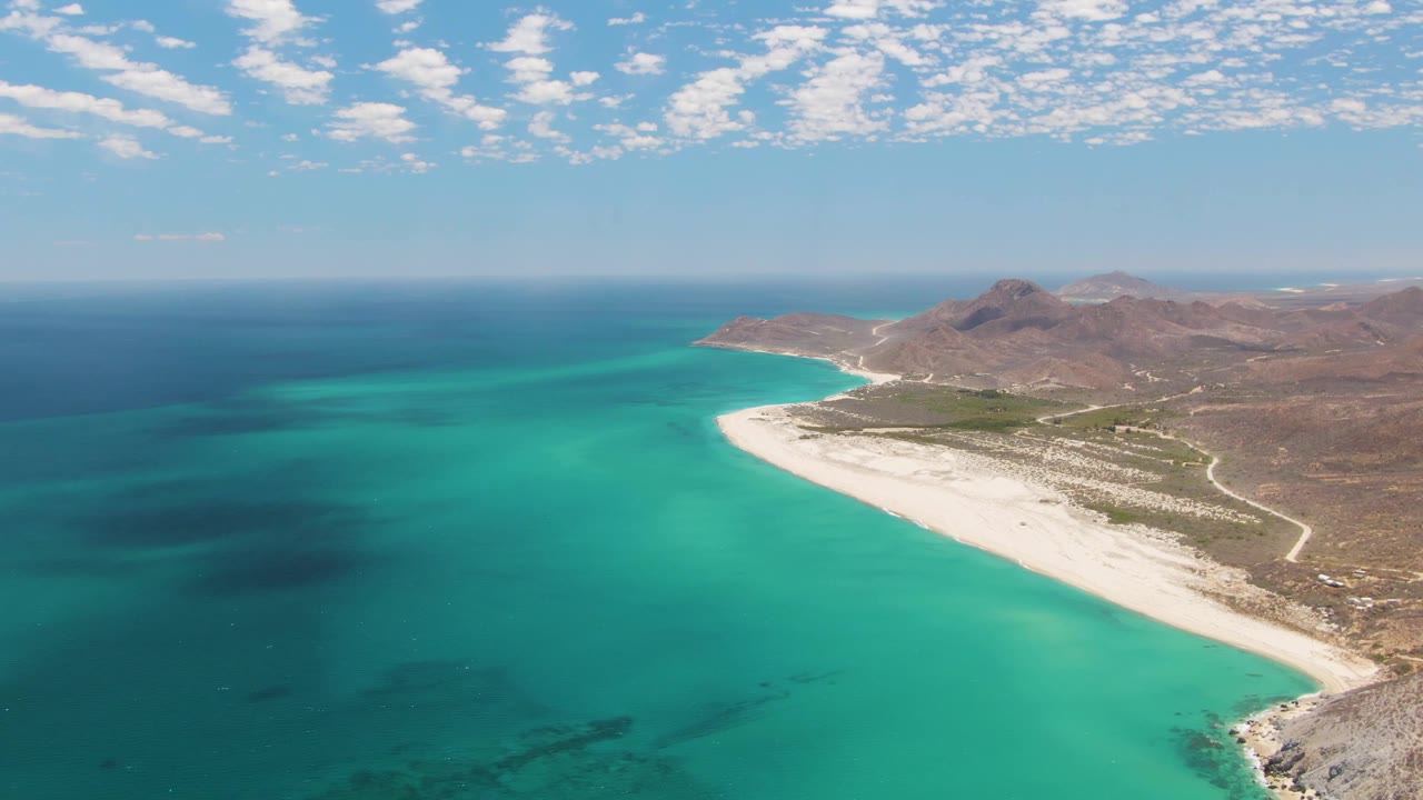 Opening aerial frame over Barakas Beach in Baja California Sur, with turquoise sea in the foreground, a pale curving beach, rocky hills inland, scattered clouds, and no visible people.