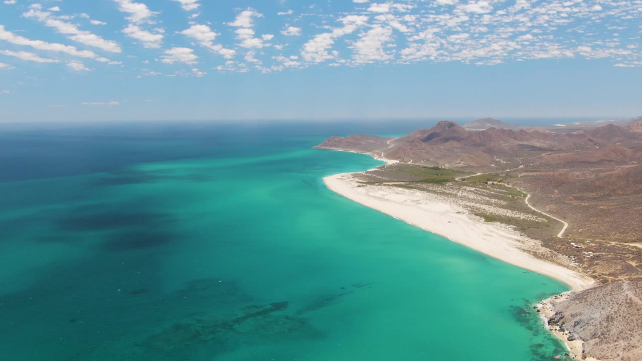Aerial view one second into the clip at Barakas Beach, showing the turquoise bay spread across the lower frame, the white sand shore to the right, rocky inland slopes, and no visible people.
