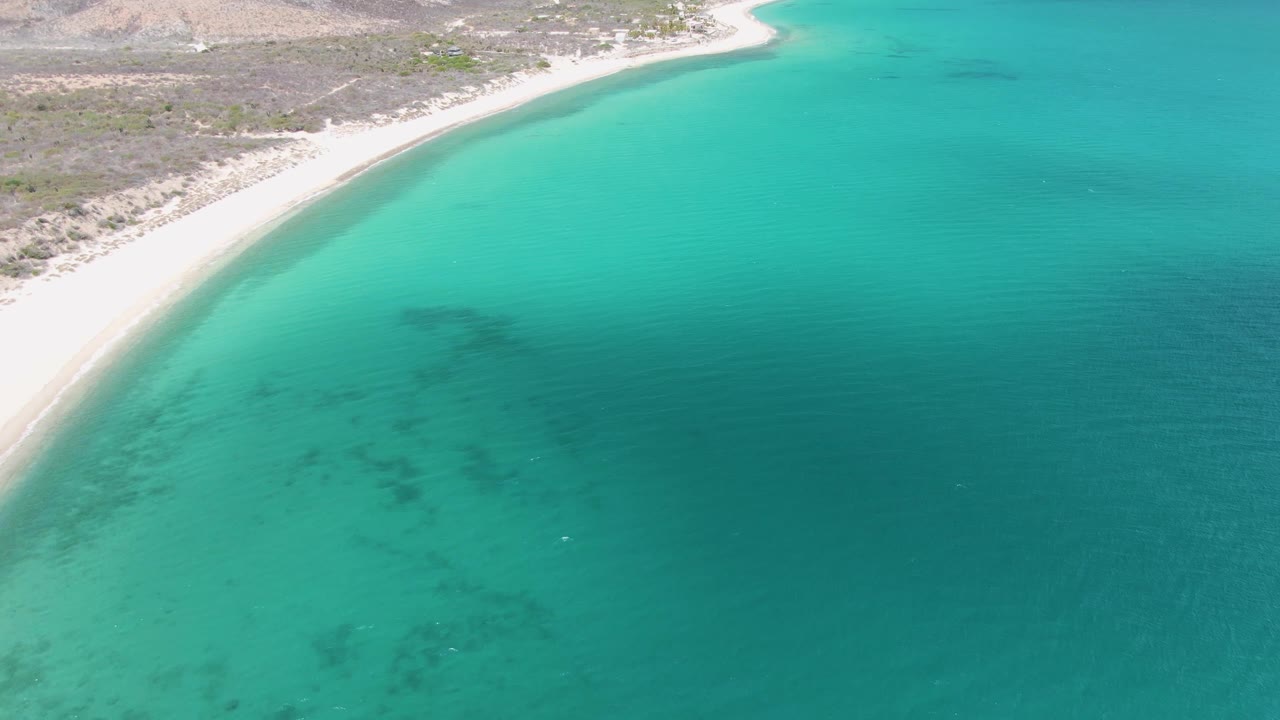 Aerial view over Barakas Beach in Baja California Sur, with clear turquoise water, pale sand, rocky dry terrain inland, and no visible people.
