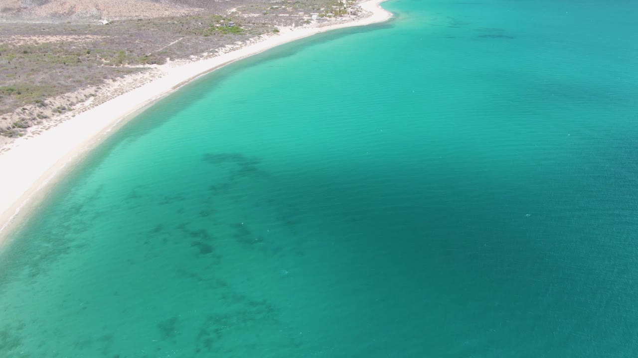 Aerial frame over Barakas Beach showing pale sand along the upper left, turquoise water across the frame, darker submerged patches, and no visible people.