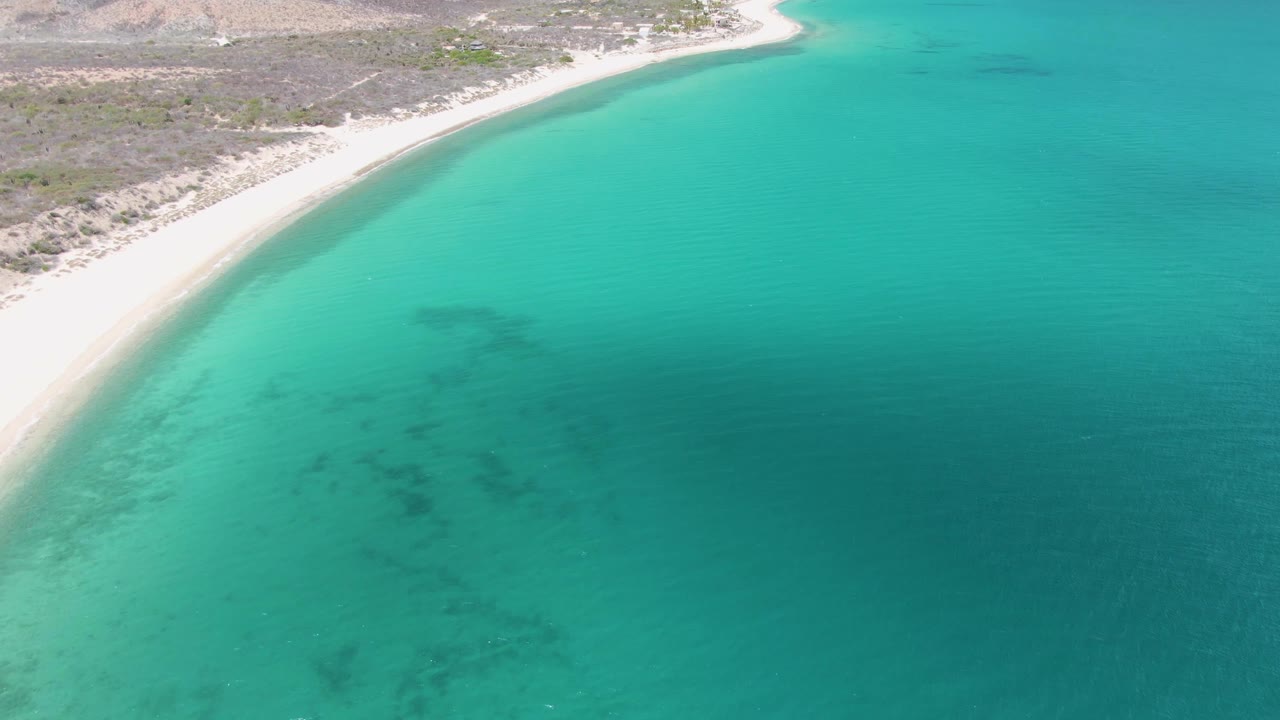 High aerial view at Barakas Beach with the shoreline running across the top of frame, open turquoise sea below, faint ripples, and no visible people.