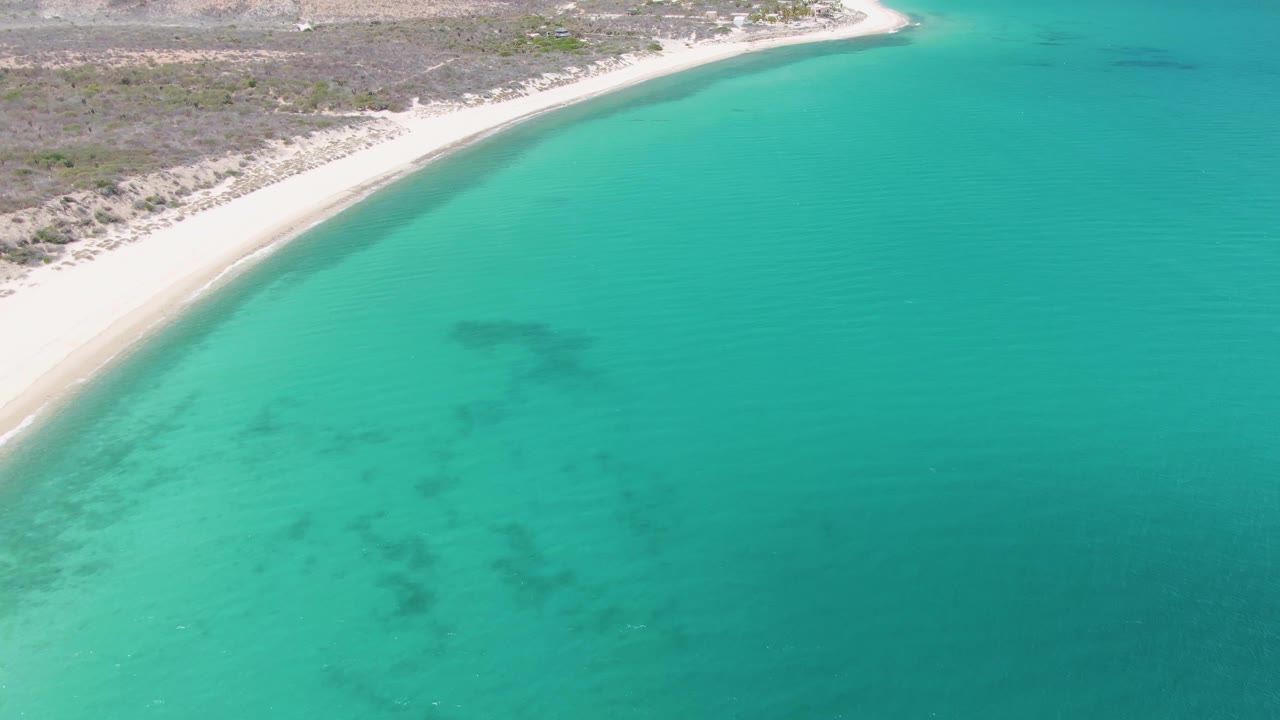 Aerial view of Barakas Beach in Baja California Sur, with a pale sand curve, dry scrub beyond the shore, turquoise water, and no clearly visible people in this first frame.