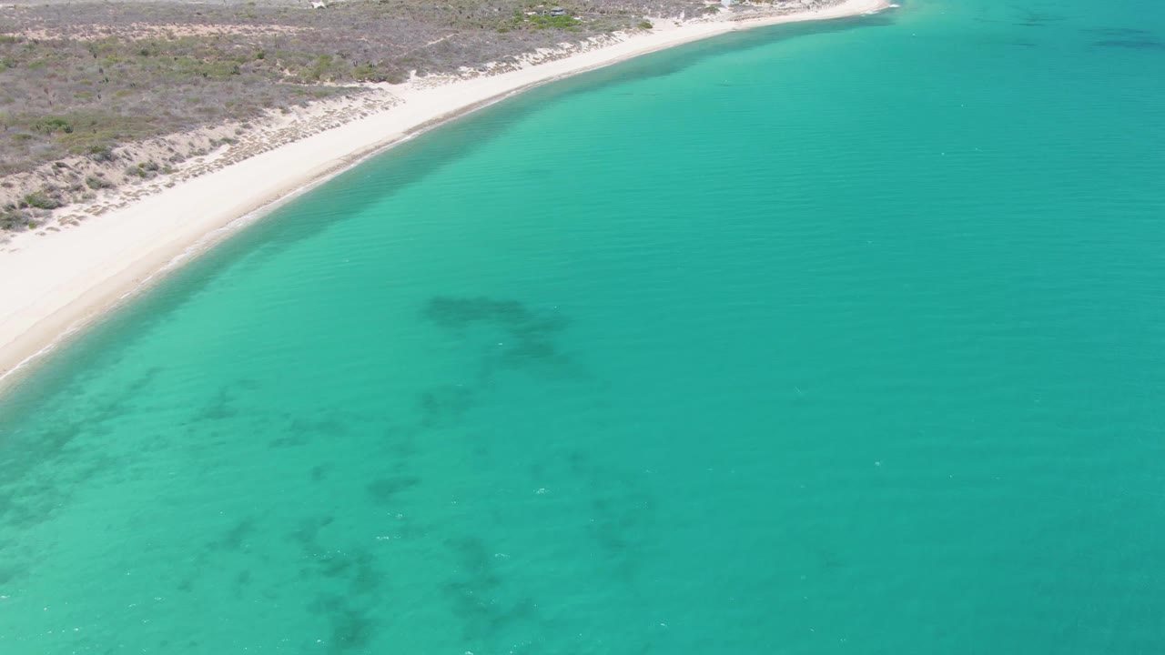 Aerial frame from Barakas Beach with a narrow pale shore, dry vegetation behind it, turquoise shallows, darker underwater areas, and tiny beach visitors beginning to appear along the sand.