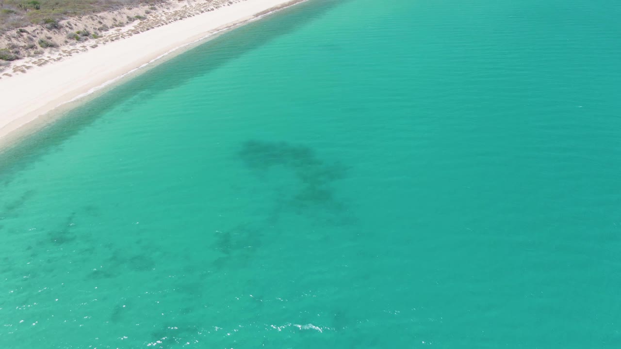 Aerial frame over Barakas Beach in Baja California Sur, Mexico, with pale sand at the top edge, turquoise water below, a dark submerged patch near center, and no visible people.