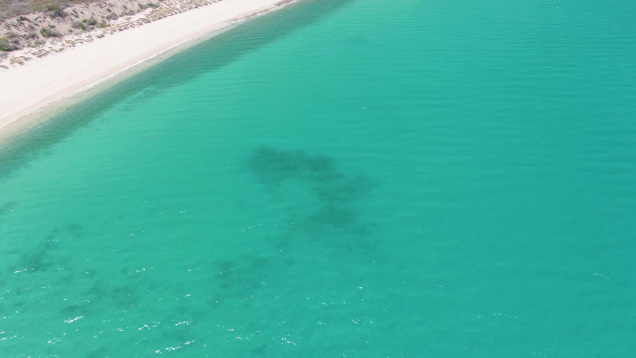 Overhead view at Barakas Beach with the white sand shoreline sliding across the upper left, bright turquoise water filling the frame, a dark underwater formation near center, and no visible people.