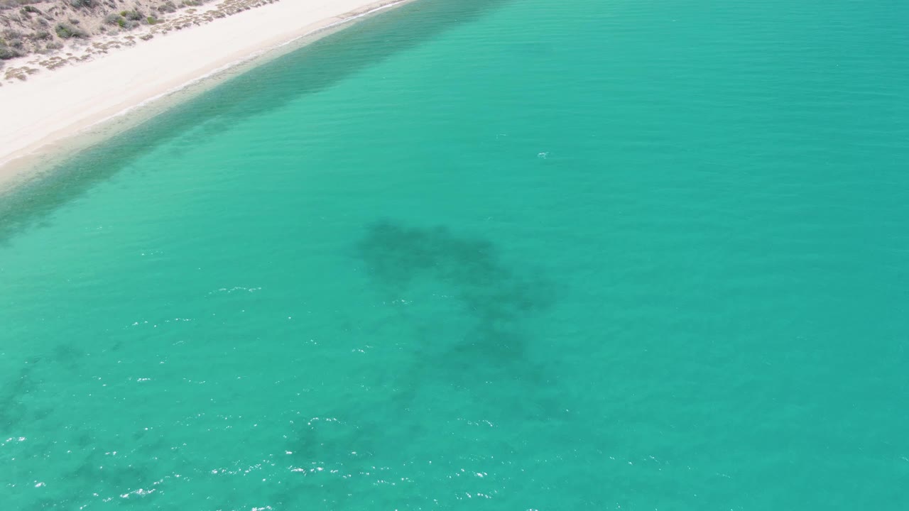Aerial view of Barakas Beach, Mexico, with a narrow band of pale beach along the top, broad turquoise ocean below, darker submerged formations under the surface, and no visible people.