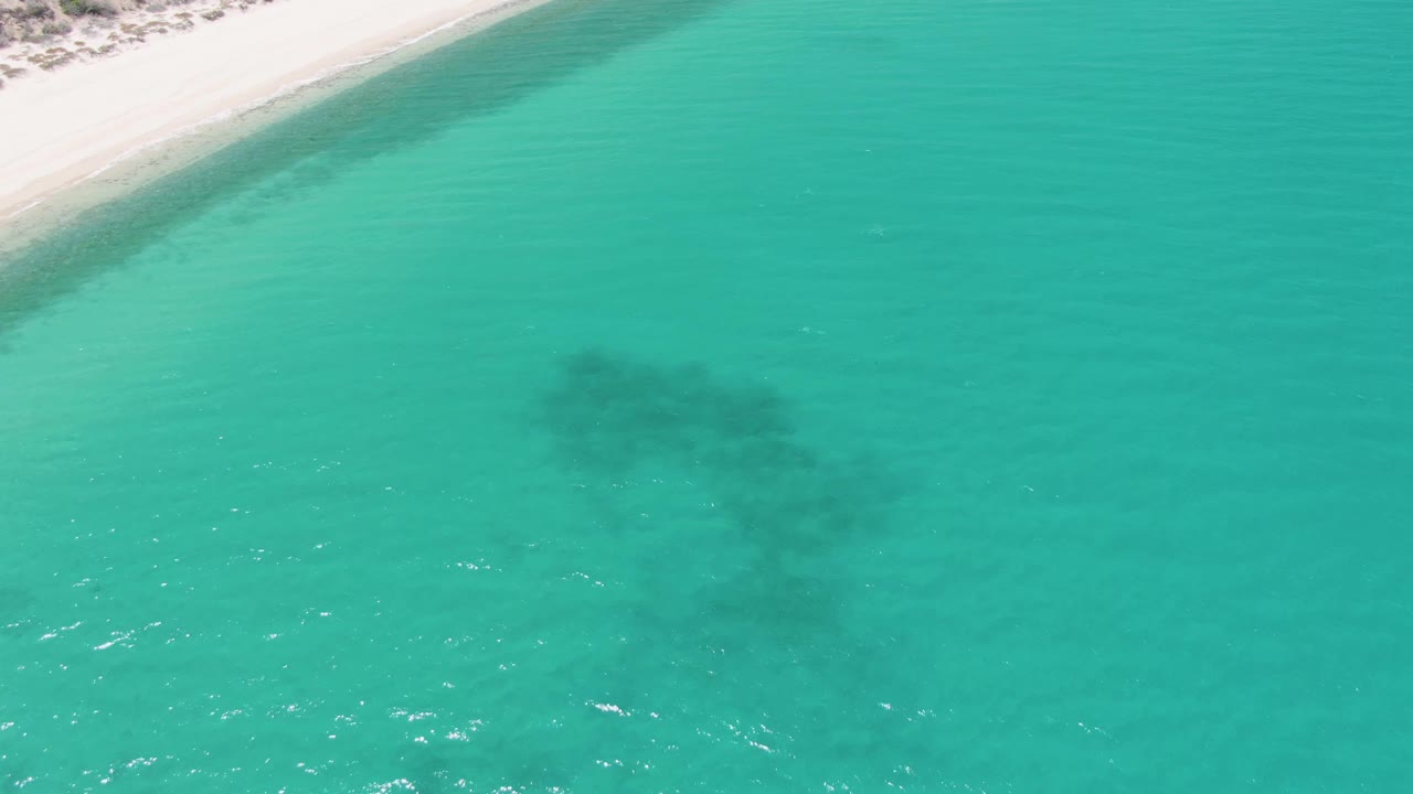 Mid-clip aerial frame at Barakas Beach showing the shoreline cropped into the upper left corner, open turquoise water across the frame, a dark submerged patch lower center, and no visible people.
