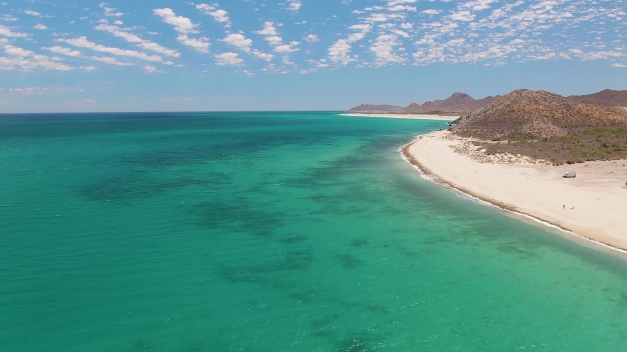 At 0:00, an aerial view over Barakas Beach shows turquoise water in the foreground, a white sand curve at right, rocky hills beyond, and tiny beach walkers visible near the shore.