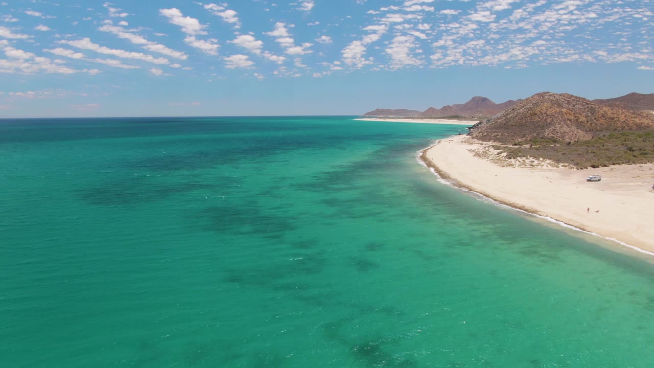 Turquoise water and curving white beach seen from above with rocky hills behind.