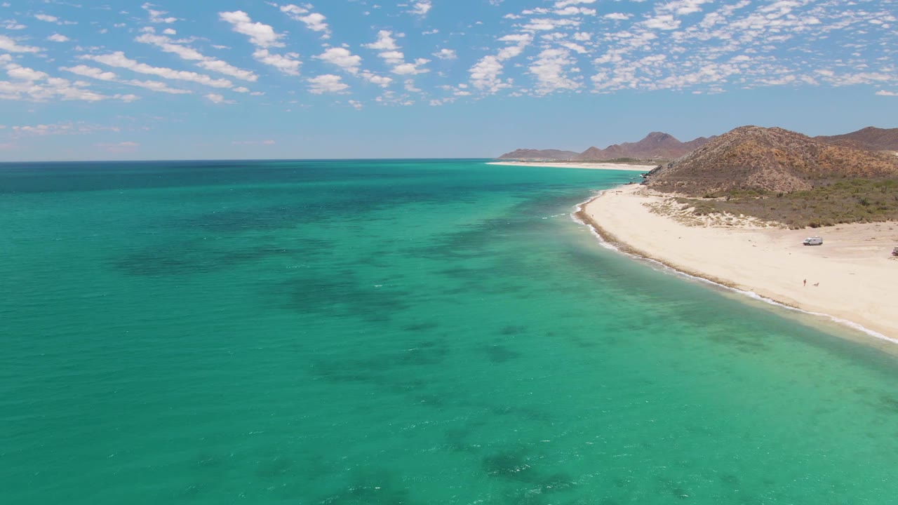 At 0:02, Barakas Beach appears with clear turquoise shallows in the lower frame, a pale sand strip curving along the right, tiny walkers, and brown hills under a patchy cloud sky.