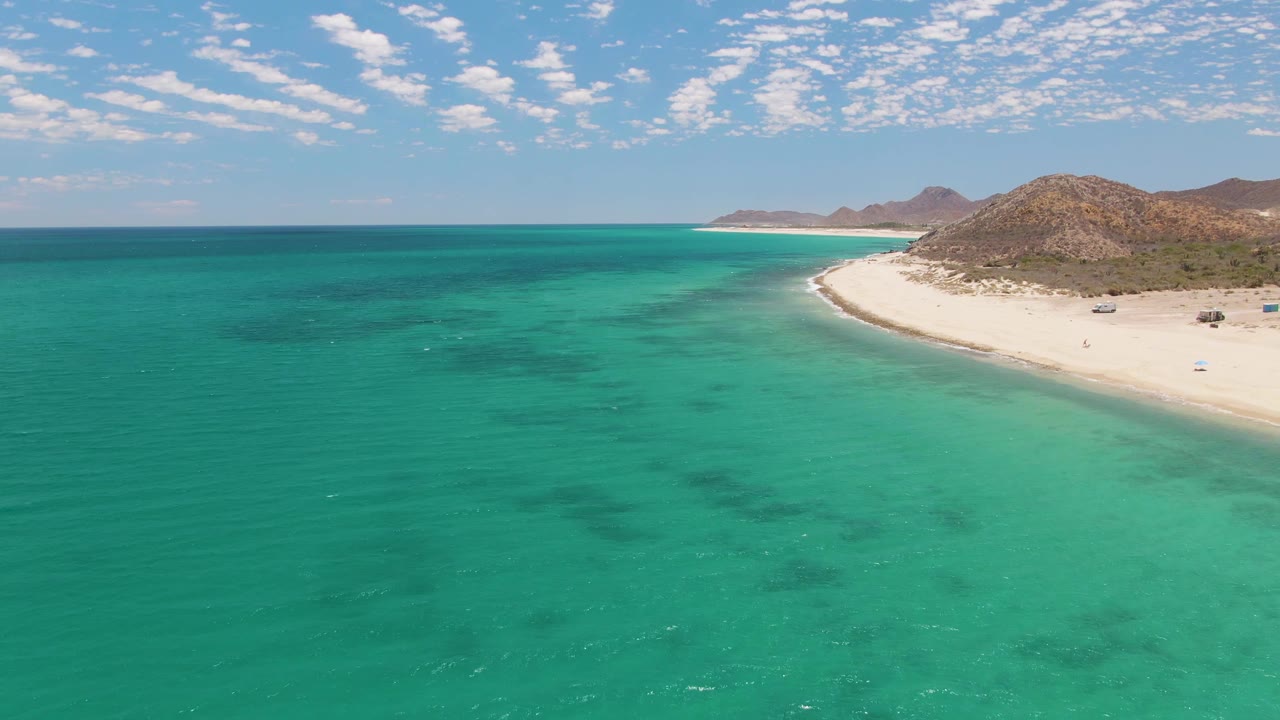 Aerial frame over Barakas Beach in Baja California Sur, Mexico, showing turquoise water, a pale curved shoreline, rocky hills, scattered clouds, and no visible people.