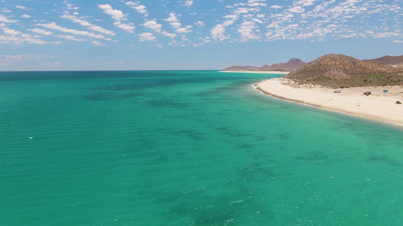 Barakas Beach appears from above with green-blue ocean texture across most of the frame, a bright sandy point at right, dry rocky hills beyond, and no visible people.