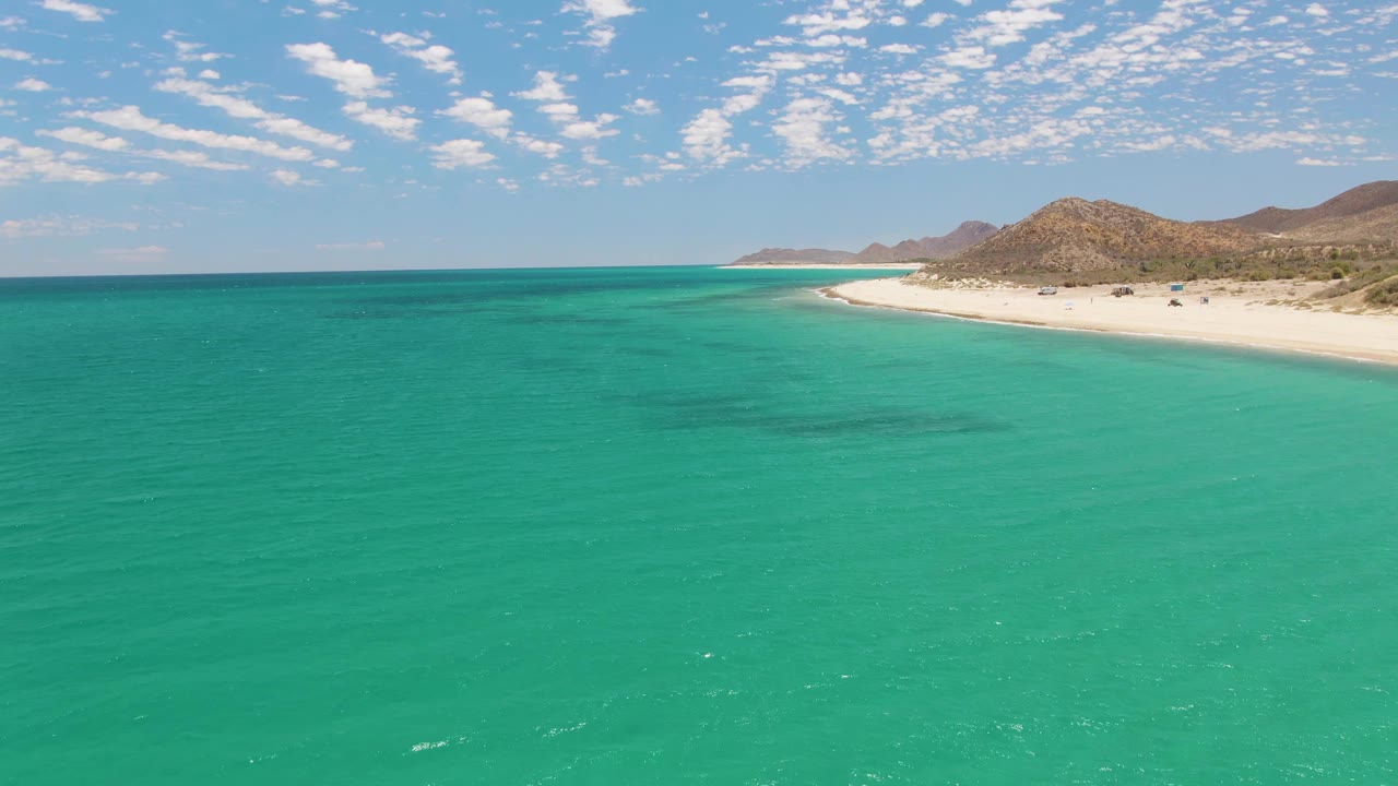 Opening frame of Barakas Beach in Baja California Sur, with turquoise water in the foreground, a pale curved shoreline at right, rocky hills beyond, scattered clouds overhead, and no visible people.