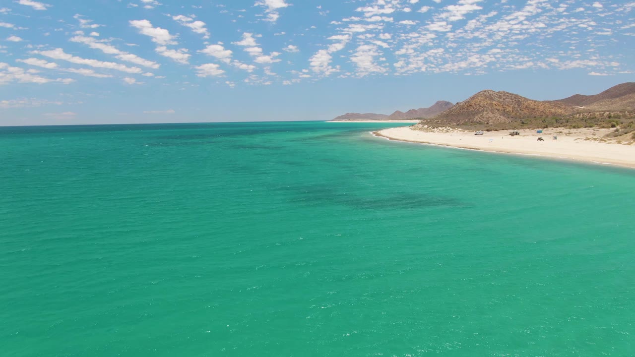 One second into the Barakas Beach clip, the drone view holds on clear turquoise water, the white beach along the right edge, brown hills in the distance, light cloud cover, and no visible people.