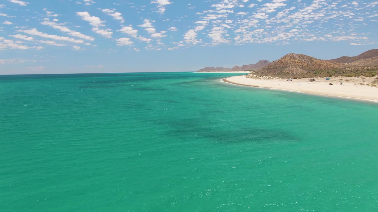 Wide ocean foreground with white beach, low hills, and scattered clouds.