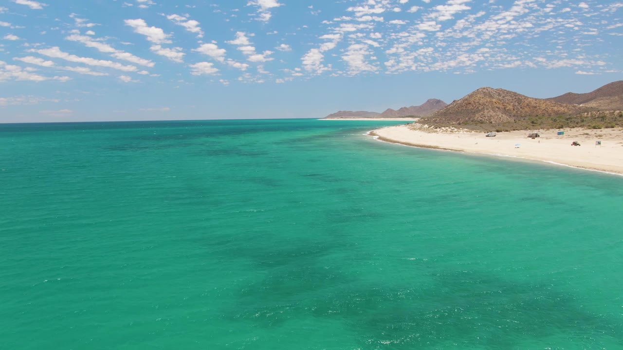 Aerial frame at Barakas Beach, Baja California Sur, showing turquoise ocean in the foreground, a white beach curving right, low brown hills beyond, scattered clouds, and tiny beach activity on shore.