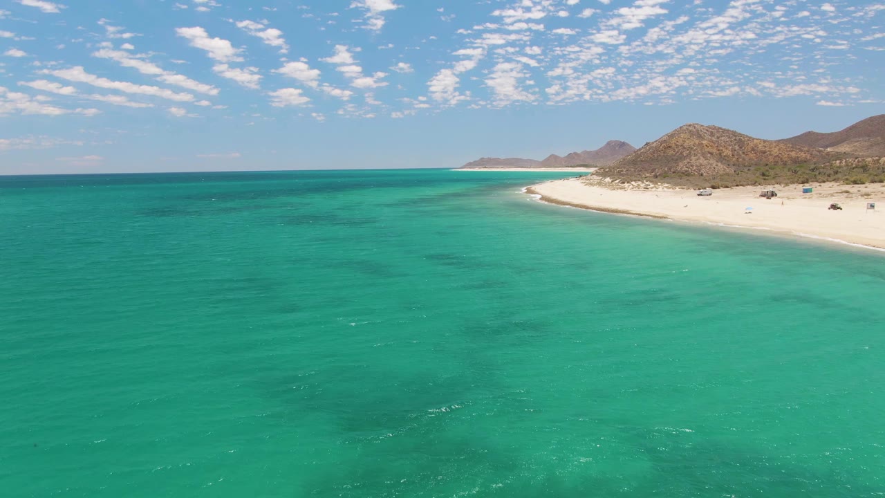 Wide drone view over calm turquoise water toward a sandy beach and hills.