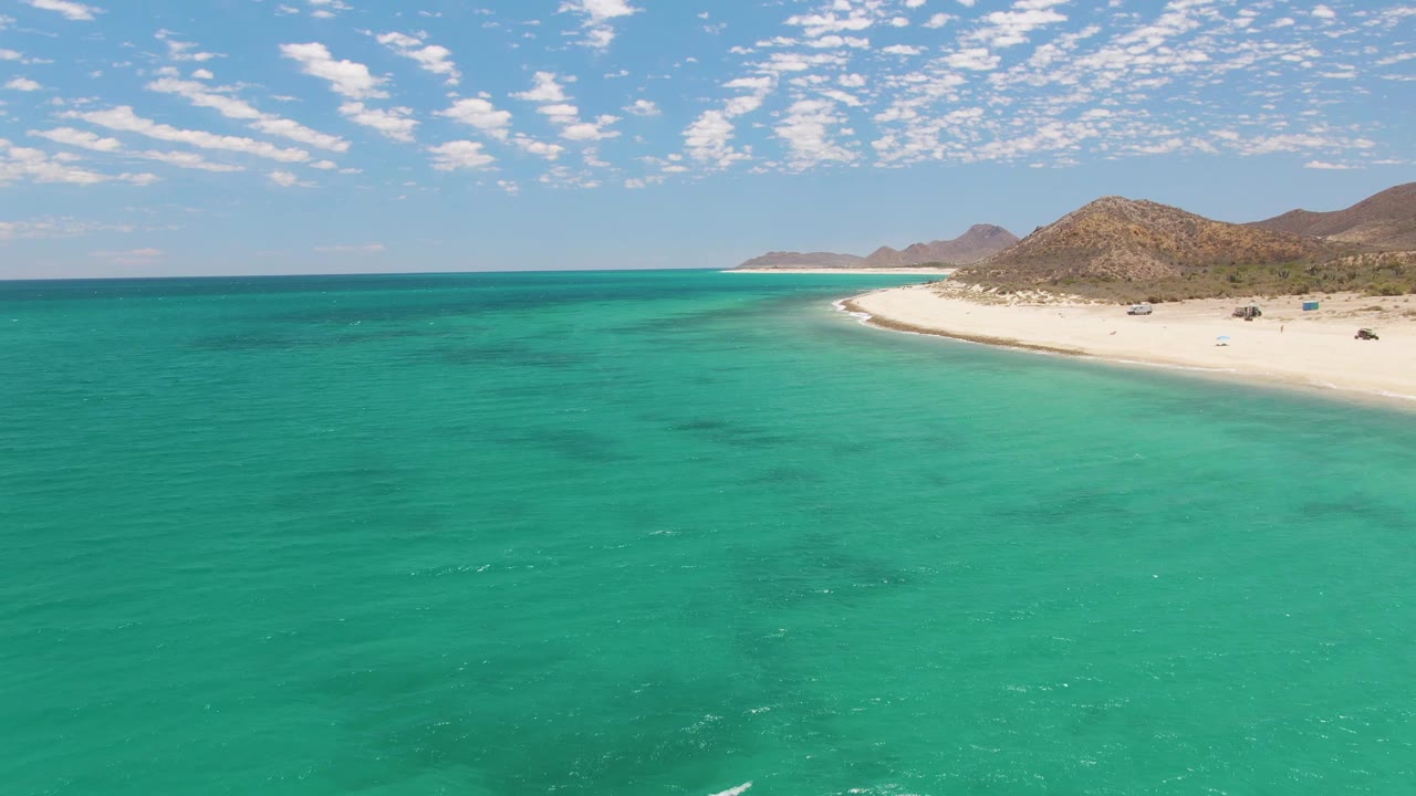 At 0:02, Barakas Beach remains on the right side of the aerial frame while broad turquoise water dominates the foreground and small beachgoers and vehicles appear as tiny details near the sand.