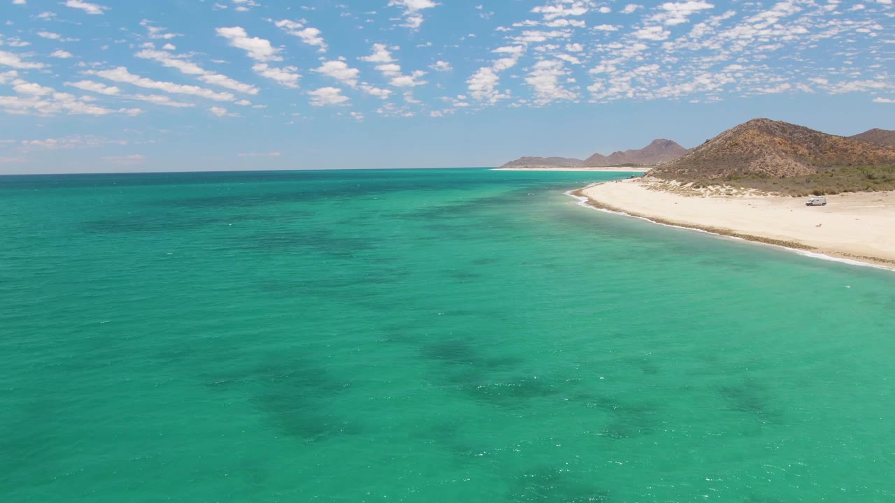 Barakas Beach aerial frame with broad clear turquoise water in front, the sandy shoreline bending along the right edge, low brown hills behind the beach, small vehicles on the sand, and no visible people.