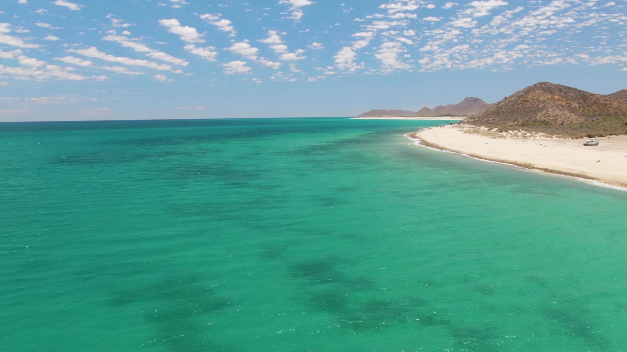 Mid-clip aerial view at Barakas Beach showing a larger expanse of turquoise water, darker patches beneath the surface, a narrow white shoreline on the right, hills in the background, and no visible people.