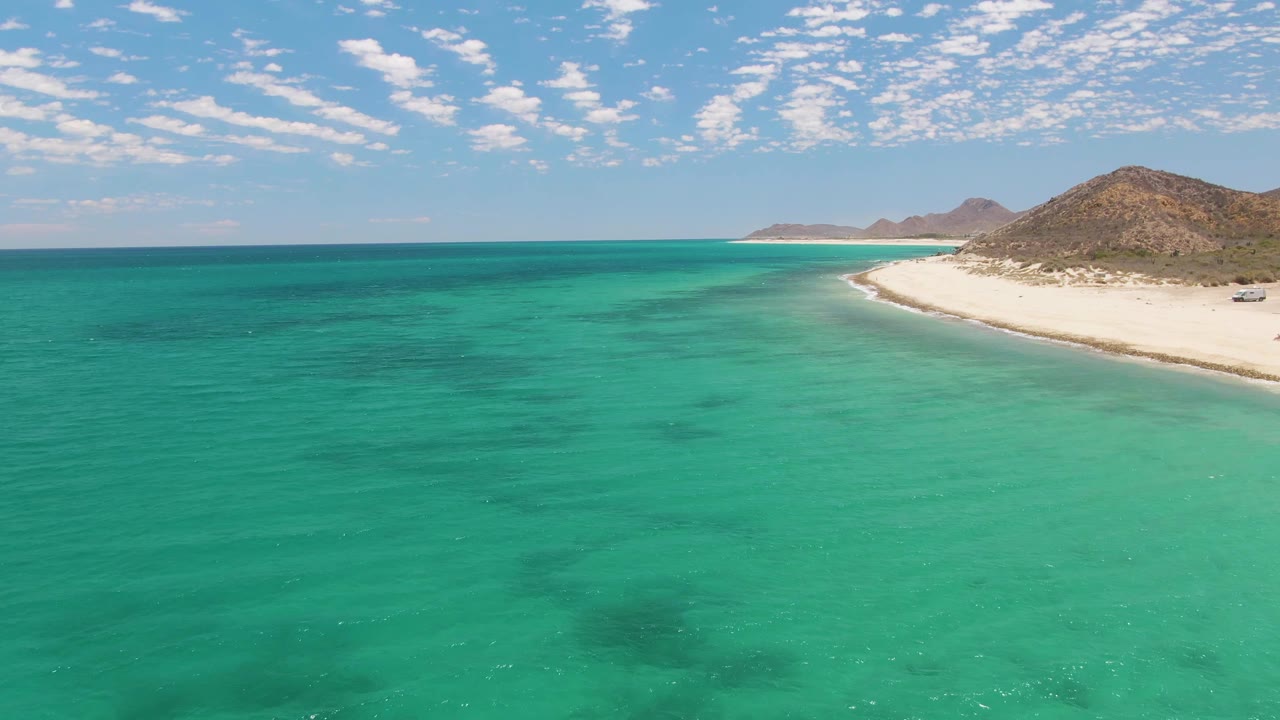 Later aerial frame of Barakas Beach with green-blue water ripples in the foreground, the pale sand strip and vehicle tracks along the right, brown hills behind, scattered cloud cover, and no visible people.