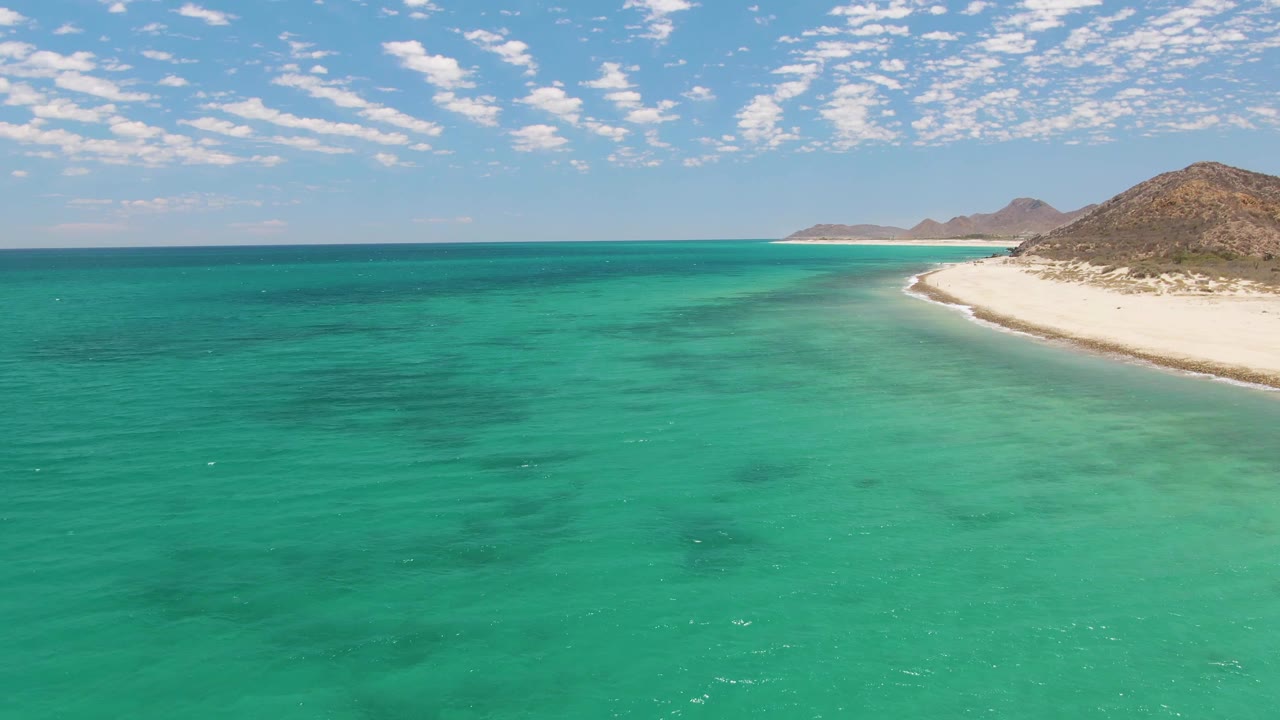 Opening frame of Barakas Beach in Baja California Sur with turquoise water in the foreground, white sand curving at right, low hills beyond, scattered clouds, and no visible people.