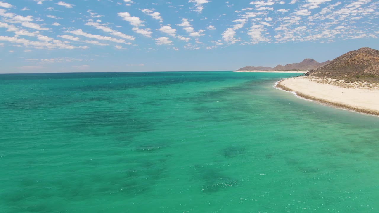 At 0:01, Barakas Beach remains framed from above with more open turquoise water across the center, the pale shoreline on the right, distant hills, and no visible people.