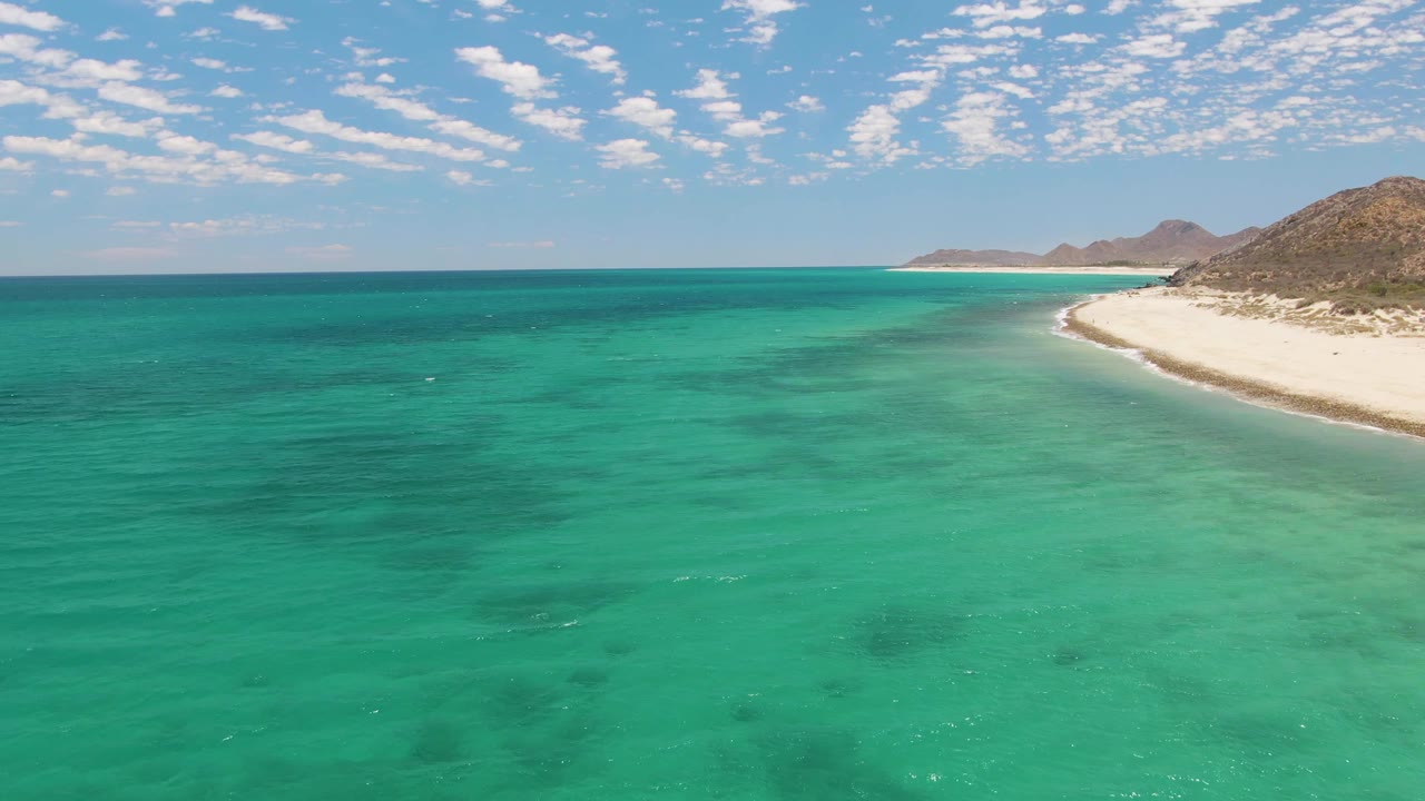 At 0:02, the elevated view over Barakas Beach shows darker patches under the turquoise water, the white beach narrowing into the distance, low hills, clouds, and no visible people.