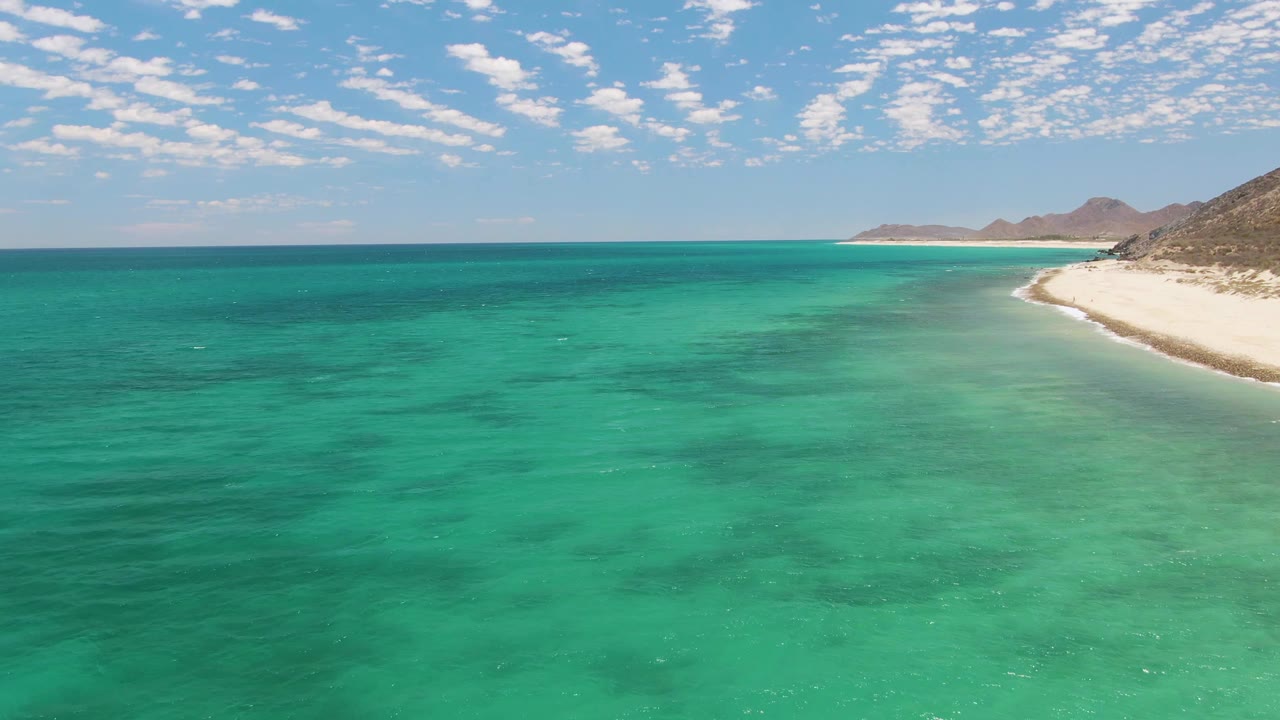 Aerial view of clear turquoise water beside Barakas Beach in Baja California Sur, with white sand and brown hills on the right and no visible people.