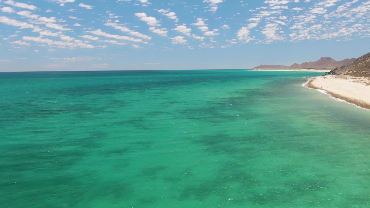 Aerial frame over Barakas Beach showing transparent green-blue water, a narrow white shoreline on the right, distant low hills, scattered cloud cover, and no visible people.